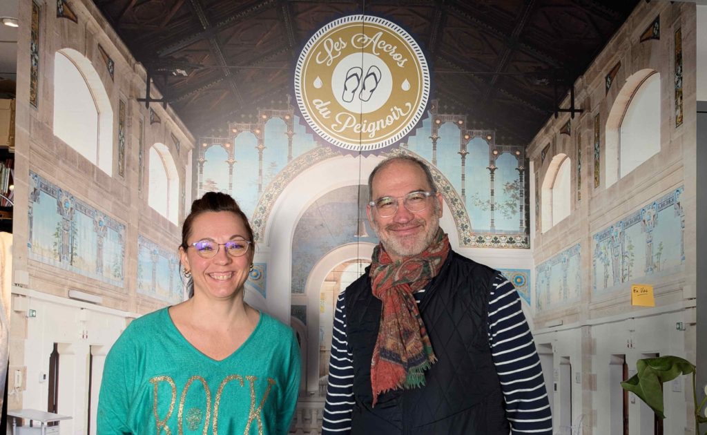 Marion et Eric posant devant l'affiche des Accros du peignoir avec son décor de thermes en trompe-l'oeil